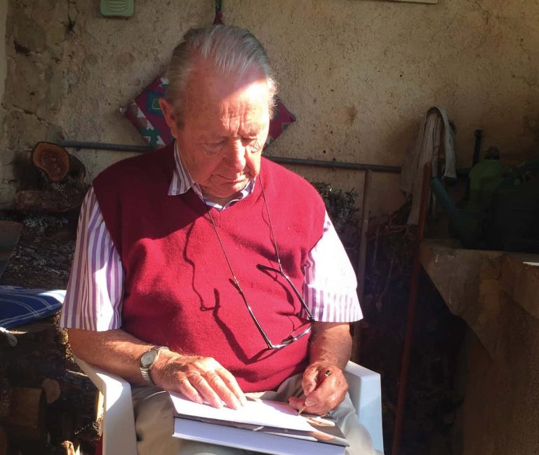 An older man wearing a red vest sits in a chair drawing.