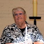An older woman with short gray hair holds a candle and looks pensive