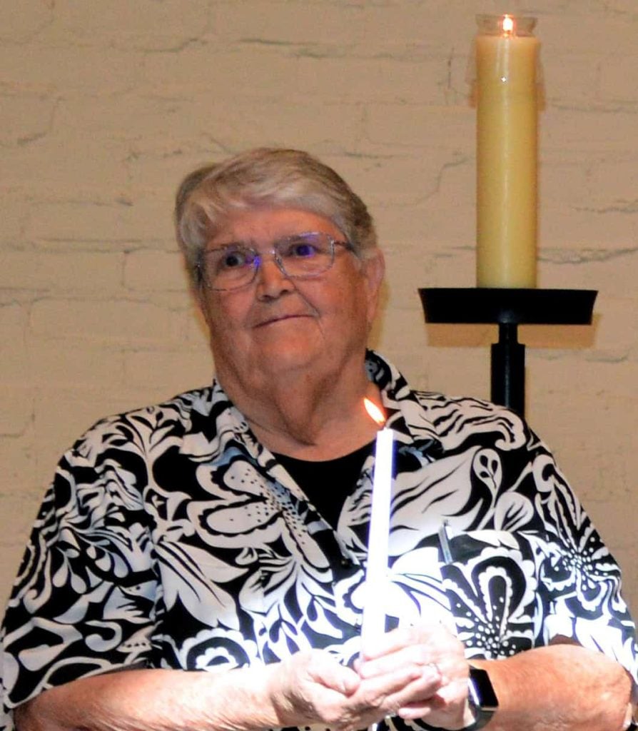 An older woman with short gray hair holds a candle and looks pensive