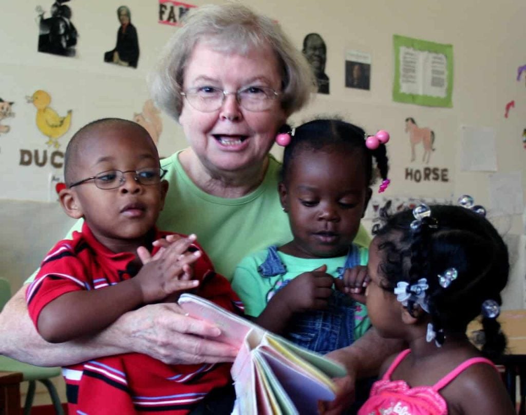 An older white woman wearing glasses reads to three Black toddlers