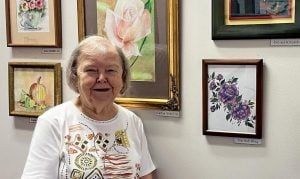 An older woman stands in front of paintings hanging on a wall