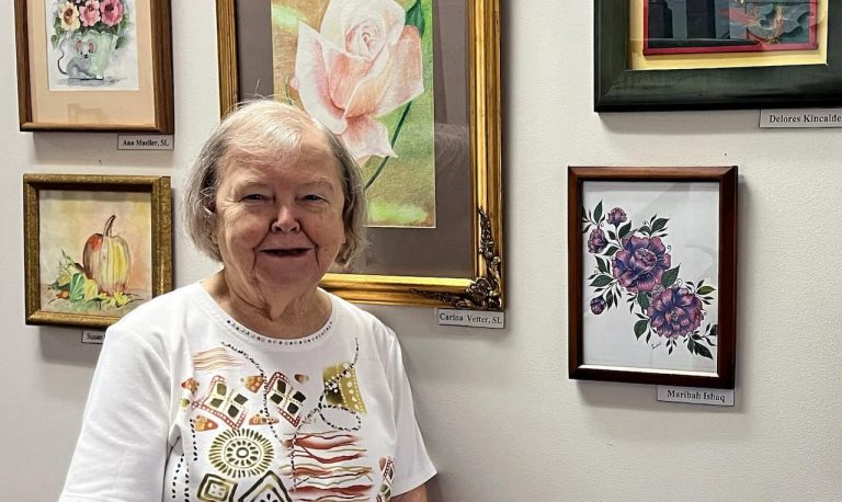 An older woman stands in front of paintings hanging on a wall