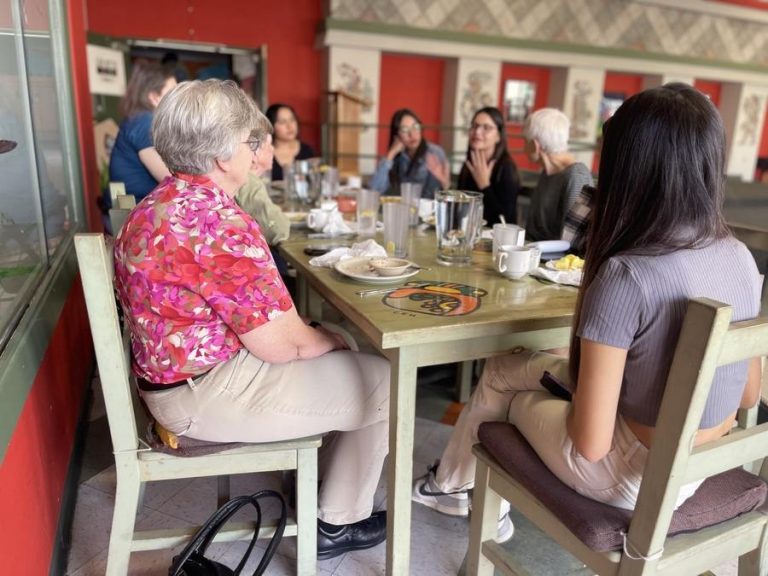 A group of people share a meal at a table together, listening to one person animatedly talk.