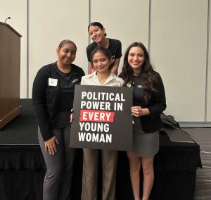 Four women smiling together in a group near a stage holding a sign that says "political power in every young woman."