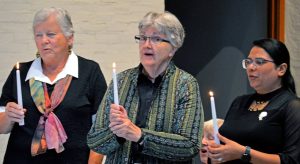 Two older women with gray hair and a younger woman with black hair and glasses hold candles