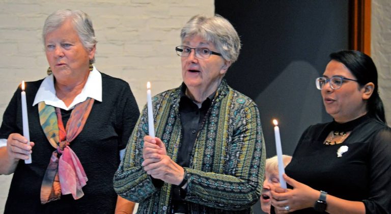 Two older women with gray hair and a younger woman with black hair and glasses hold candles