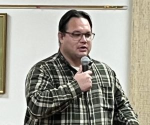 A man wearing glasses and a flannel shirt speaking into a microphone indoors.