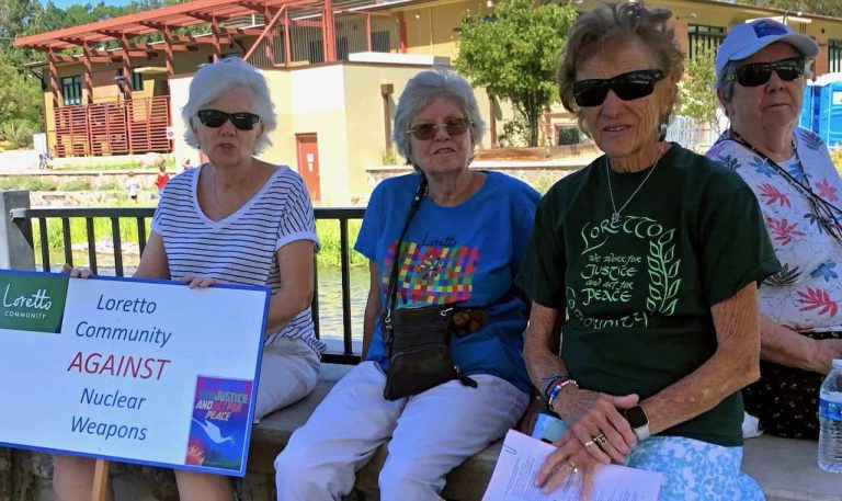 Four older women sit outdoors with an anti-nuclear weapons sign