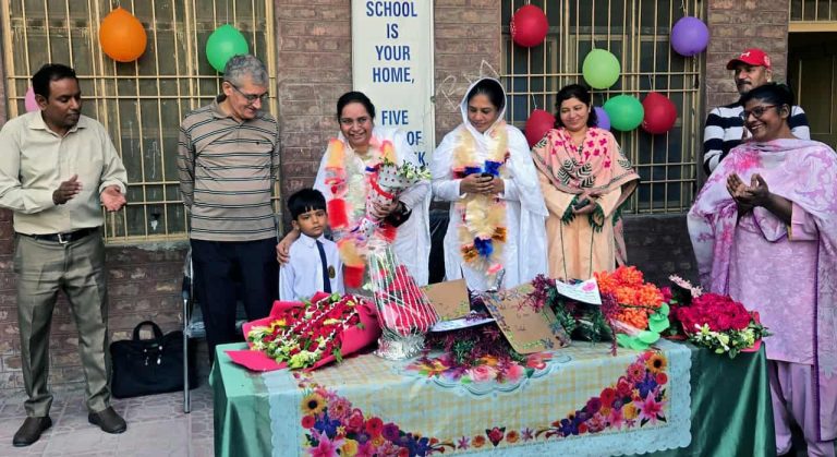 Women wearing saris and men and one young boy in a white shirt and tie celebrate outdoors in Pakistan next to a table strewn with flowers