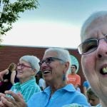 Older women smile and applaud outdoors as they watch a performance.