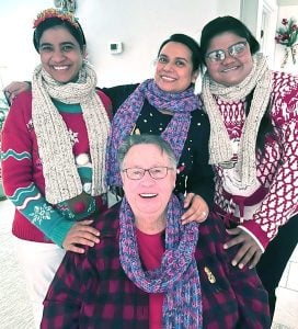 Three women standing and one woman sitting smiling together for a group picture indoors while wearing warm sweaters and scarves.