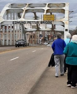 Three women walking on the sidewalk of a road bridge with a large white arch and the sign Edmund Pettus Bridge on an overcast day.