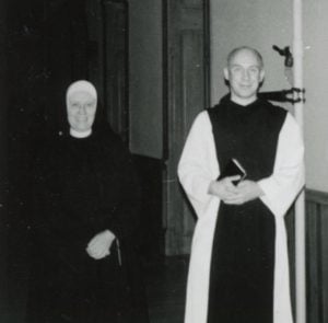 Black and white archival photo of a habited nun standing for a photo with a monk in the hallway.