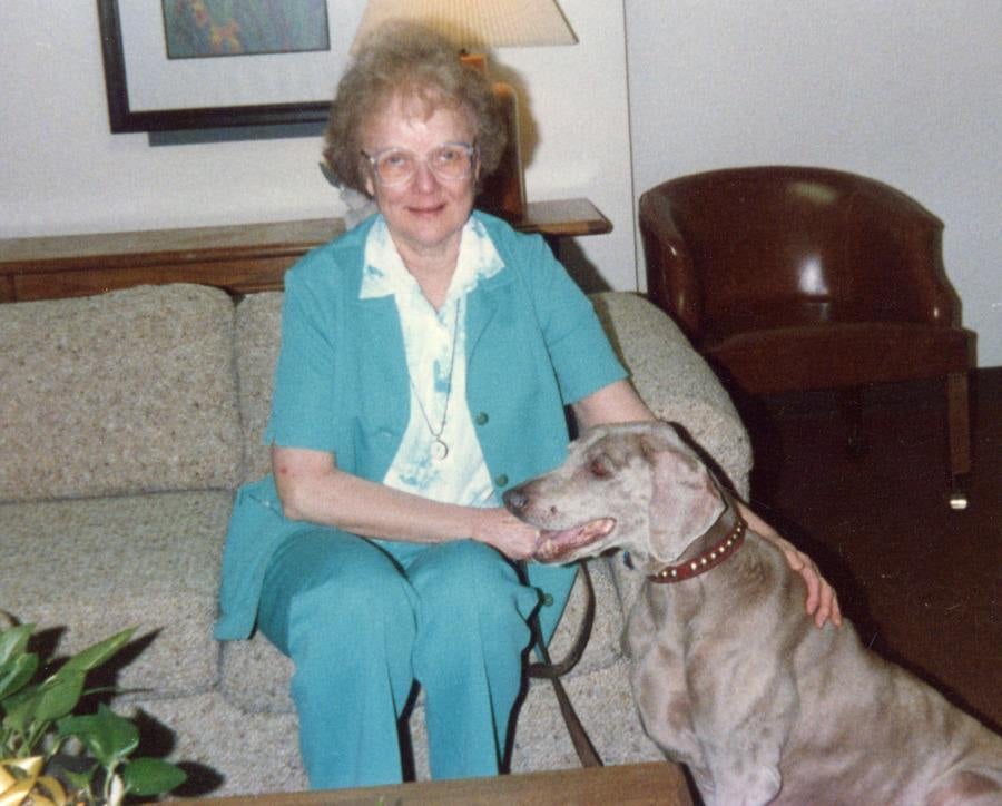 A color archival photo of a woman in a light blue pantsuit sitting on a living room couch, holding the leash of the large, short-haired dog sitting next to her on the floor.