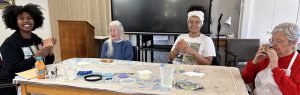 Four women smiling together while sitting at a craft table painting small wooden boxes inside.