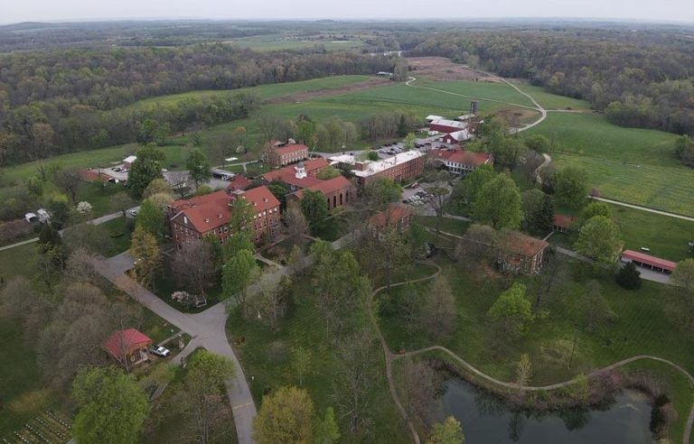 An aerial view of the Loretto Motherhouse, with multiple red brick buildings in the center surrounded by trees, fields, a pond, paths, and a circular road.