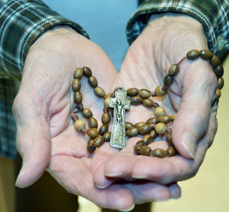A rosary made of carved wooden beads and a metal cross on a cord is gently cupped in the hands of an older person.