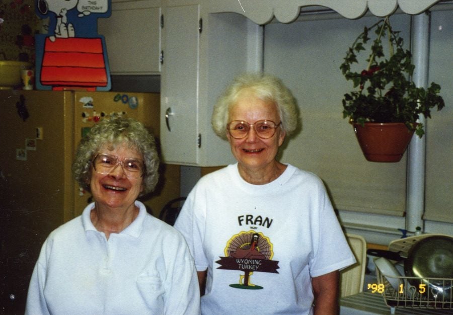 Archival color photo of two women posing in a kitchen for a photo. In the background, a large Snoopy decoration sits on top of the refrigerator.