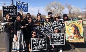 A group of protestors smiling together for a group picture while holding up their signs, many of which say, "Protect Oak Flat Apache Stronghold." They are standing outside on a sunny day.