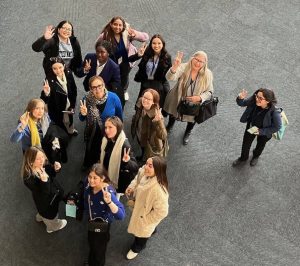 A group of women of various ages holding up peace signs for a group photo. The photo is taken from above looking down on the group.