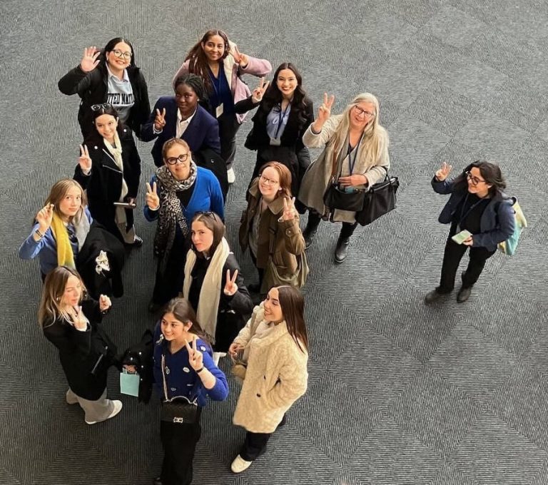 A group of women of various ages holding up peace signs for a group photo. The photo is taken from above looking down on the group.