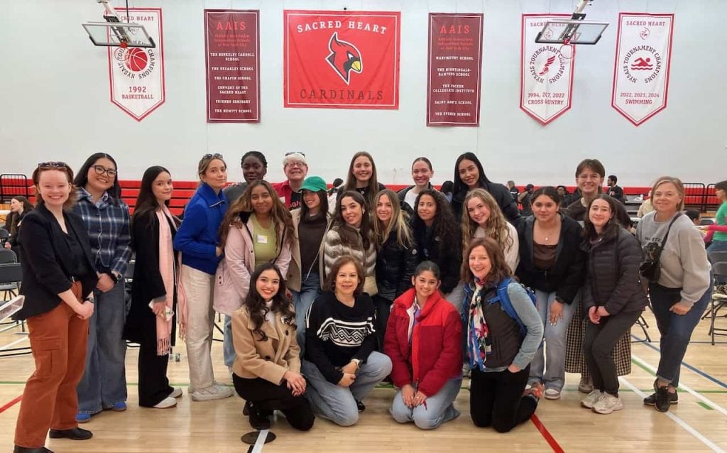 A group of 22 teenagers and a few adults smile for a picture in a school gym. Above them are several red banners that say "sacred heart cardinals"