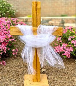 A wooden cross draped in white mesh fabric stands in front of pink flowering bushes.
