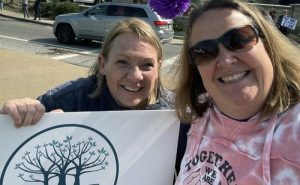 Two women, one wearing sunglasses and one holding a large sign with a tree design, smile for a selfie together outdoors on a sunny day.