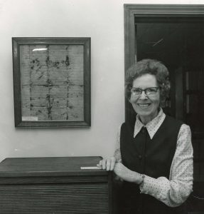 Archival black and white photo of a woman in glasses standing next to a framed historical document.