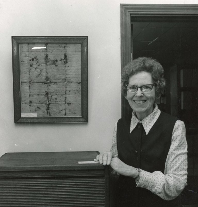 Archival black and white photo of a woman in glasses standing next to a framed historical document.
