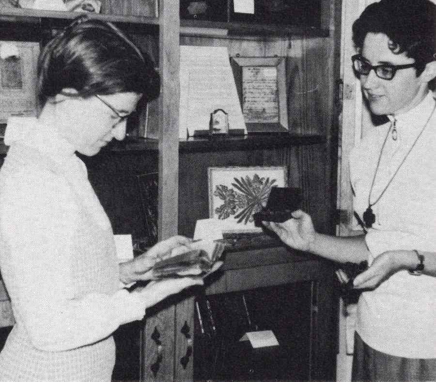 Archival black and white photo of two women looking at several historical items that they are holding.