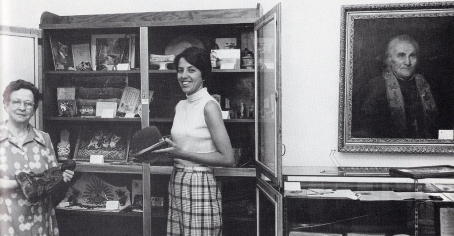 Archival black and white photo of two women standing in front of an open curio case holding objects. The older woman on the left is holding a statue of the Buddha; the younger, dark-haired woman on the right is holding a bowler-style hat.