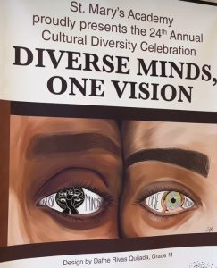 A publicity with two different faces pressed together on their temples, one brown and one white, only pictured is their eyes next to eachother. The publication says, "St. Mary's Academy proudly presents the 24th Annual Cultural Diversity Celebration DIVERSE MINDS, ONE VISION Design by Dafne Rivas Quijada, Grade 11