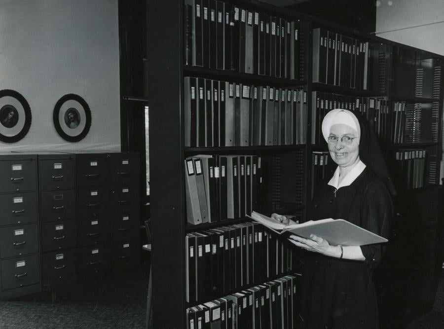 Archival black and white photo of a habited nun standing in front of a bookshelf holding an open book.