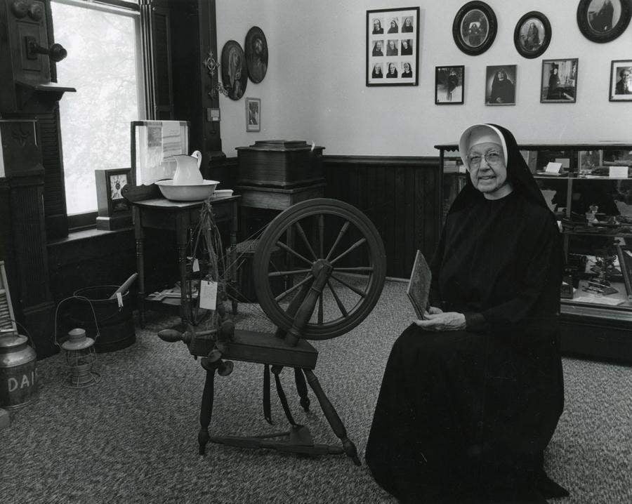 Archival black and white photo of an older nun in a habit sitting next to an old spinning wheel in a room displaying many artifacts.