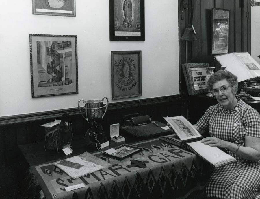 Archival black and white photo of a woman sitting at a table displaying a number of historical items.