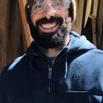 A white man with a dark beard smiling outside as he wears clear working goggles, a tan beanie and blue sweatshirt.