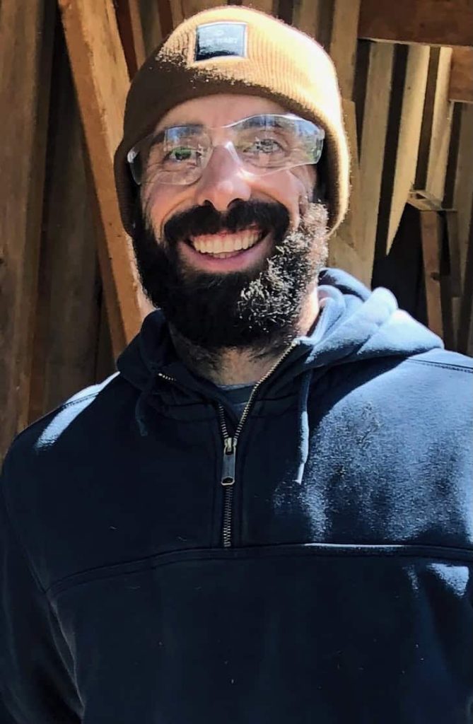 A white man with a dark beard smiling outside as he wears clear working goggles, a tan beanie and blue sweatshirt.