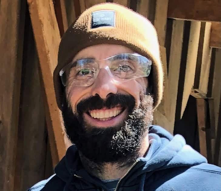 A white man with a dark beard smiling outside as he wears clear working goggles, a tan beanie and blue sweatshirt.