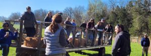 A dozen people are shown outside on a large wagon on a bright sunny day listening to a farmer discuss conservation practices.