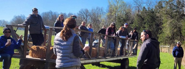 A dozen people are shown outside on a large wagon on a bright sunny day listening to a farmer discuss conservation practices.
