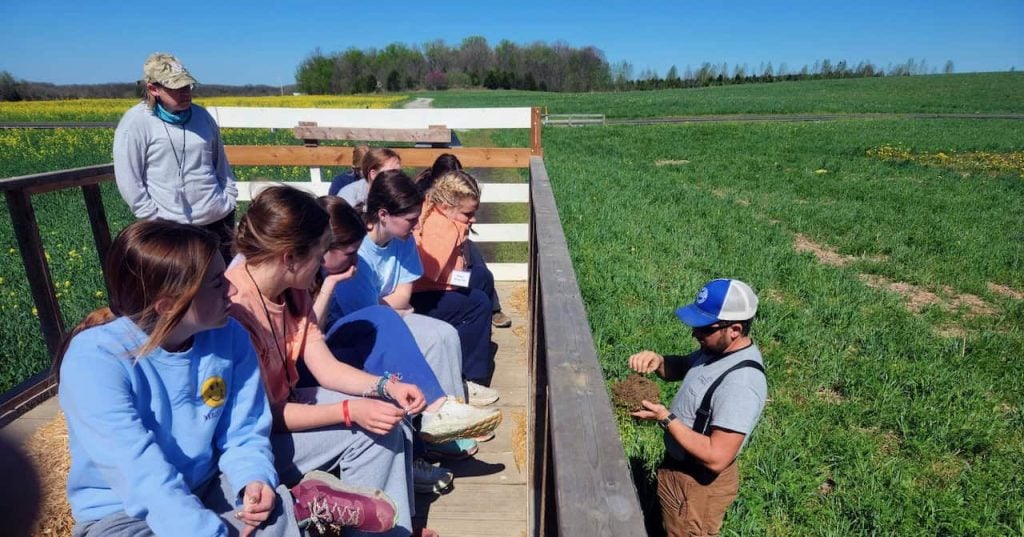 Outside, a farmer teaches a group of high schoolers about healthy soil. They are all looking intently at the farmer and very engaged.