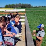Outside, a farmer teaches a group of high schoolers about healthy soil. They are all looking intently at the farmer and very engaged.