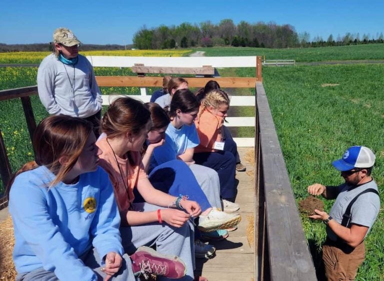 Outside, a farmer teaches a group of high schoolers about healthy soil. They are all looking intently at the farmer and very engaged.