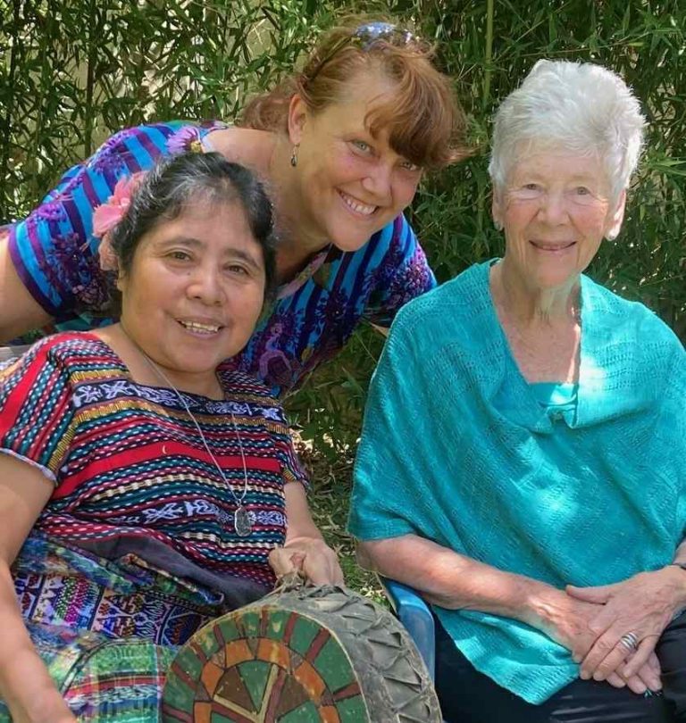 Three women, one in traditional Guatemalan patterns, lean in for a photo together in the dappled shade.