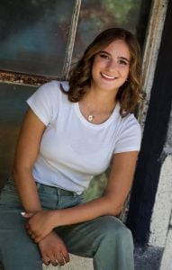A high-school aged girl smiling at the camera. She is leaning to the side and is wearing a white t-shirt and jeans.