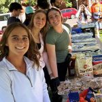 Three high school students smiling outside as they work at a lunch and clothing drive in their city.