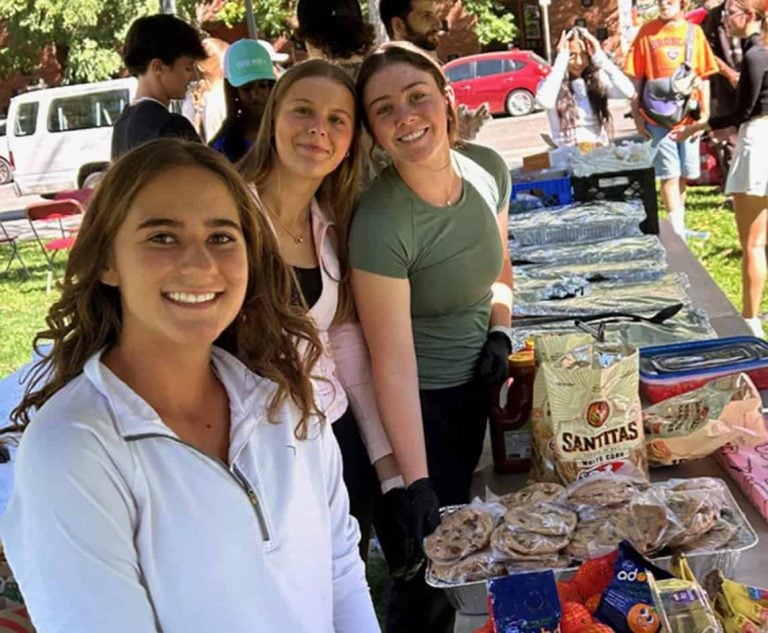 Three high school students smiling outside as they work at a lunch and clothing drive in their city.