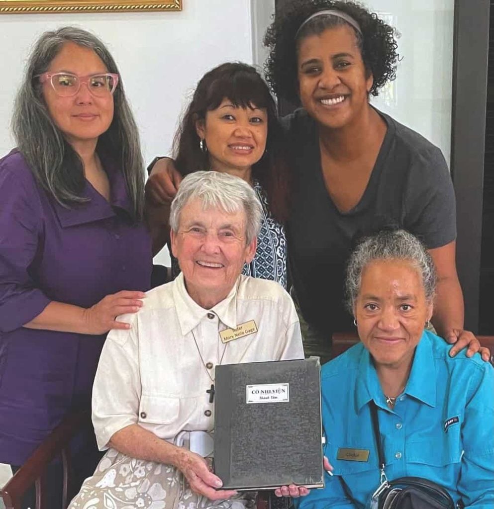 Former Vietnamese adoptees visit an orphanage that was once home. Four adoptees and a religious sister smile for a photo together as they hold a record book.