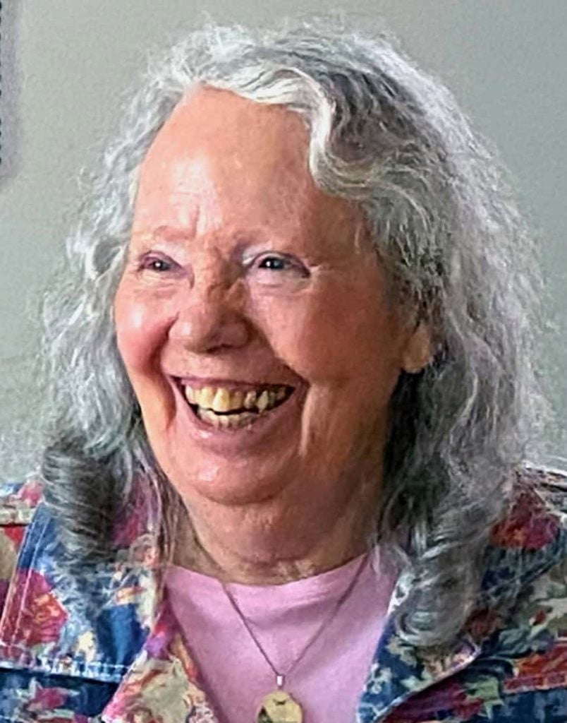 An older white woman with medium length grey hair and a pink shirt is slightly turned to the side and smiling big to somebody.